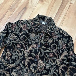 May include: A black button-down jacket with a floral tapestry design in shades of green, brown, and tan. The jacket has a collared neckline and long sleeves. The fabric has a woven texture. The jacket is a vintage piece.