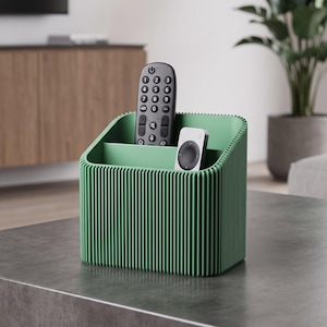 May include: A sage green desk organizer with a vertical ribbed texture and two interior compartments. The organizer holds a black television remote control with a numeric keypad and a small white circular remote control with a central button.