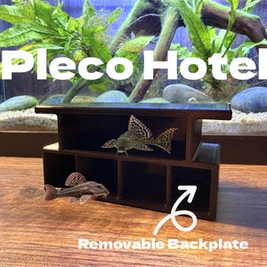 Peut inclure: Décoration d'aquarium marron foncé à compartiments multiples, intitulée "Pleco Hotel", avec une plaque arrière amovible. Deux poissons sont visibles à l'intérieur de la structure. L'arrière-plan montre un aquarium avec des plantes et des roches.