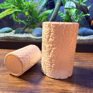 Peut inclure: Deux jardinières cylindriques blanc cassé avec un motif texturé en forme d'écorce. Une jardinière est droite, l'autre est couchée sur le côté. Les jardinières sont sur une surface en bois, avec un aquarium en arrière-plan.