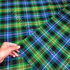 May include: A fabric featuring a vibrant plaid pattern with intersecting lines of bright green, black, deep blue, and thin yellow stripes. The textile displays a repeating tartan design across the surface, with the text Premier Apparelshop visible.
