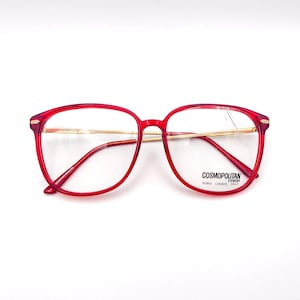 May include: Red-framed eyeglasses with gold-colored arms. The glasses have a square shape and the text "Cosmopolitan Eyewear Demo Lenses Only" is printed on the lower right lens.