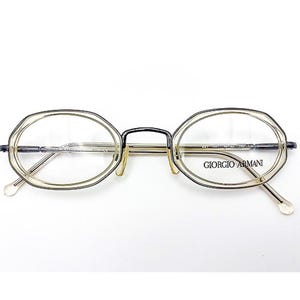 May include: Oval-shaped eyeglasses with a light-colored frame and clear lenses. The glasses have a thin, dark bridge and temples. The brand name "GIORGIO ARMANI" is printed on the right lens. The overall design is minimalist and elegant.