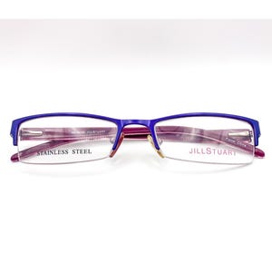 May include: Purple and blue eyeglasses with a semi-rimless design. The frames are made of stainless steel and feature the brand name "JILL STUART". The glasses have a rectangular shape and are set against a white background.