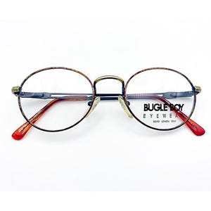May include: Oval-shaped eyeglasses with a thin, patterned brown frame and red temple tips. The bridge and hinges are gold-toned. The lenses are clear, and the brand name "BUGLE BOY EYEWEAR" is printed on the right lens.