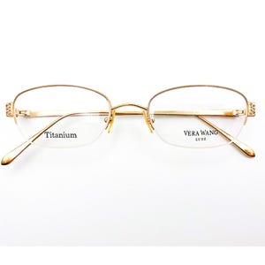 May include: Gold-toned Vera Wang Luxe eyeglasses with a half-rim design. The frames are adorned with small decorative elements on the temples. The lenses are clear, and the text "Titanium" and "Vera Wang Luxe" are visible.