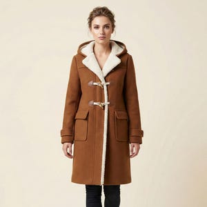 Op de afbeelding: Een bruine duffelcoat met een capuchon en knevelsluitingen. De jas heeft een crèmekleurige voering en afwerking, en twee voorzakken. Het model draagt een zwarte jeans en bruine laarzen.