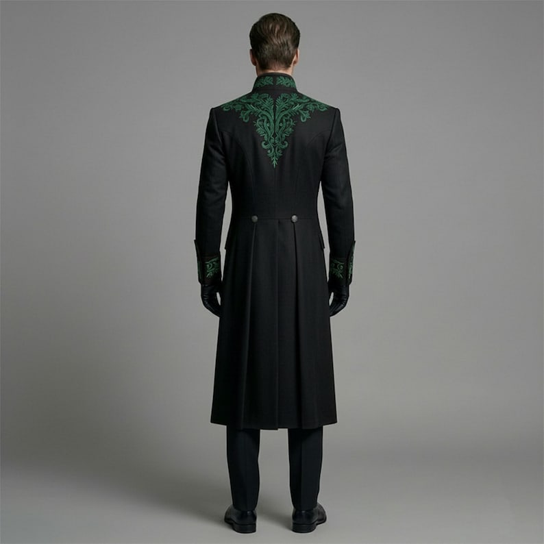 May include: A long black coat with green embroidered detailing on the shoulders and cuffs. The coat has a high collar and a button closure. The person is wearing black gloves and black pants. The coat is a formal style.