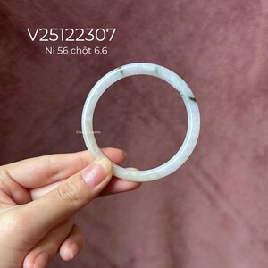 May include: A round, white jadeite bangle with subtle green veining. The text "V25122307 Ni 56 chọt 6.6" is visible. The bangle is held against a pink background. A Dreamy Gems logo is in the bottom left corner.