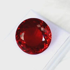 May include: A large, round, faceted gemstone in a deep red hue. The gem is displayed on a white surface, highlighting its sparkle and clarity. The cut of the stone creates many reflective surfaces, enhancing its beauty.