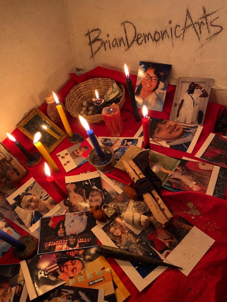 Pu&ograve; includere: Una superficie in tessuto rosso coperta da varie fotografie di persone, diverse candele accese gialle, blu, rosse e nere, un cesto intrecciato contenente piccoli oggetti, un piccolo specchio e un fascio di legno. Il testo BrianDemonicArts &egrave; scritto sulla parete.