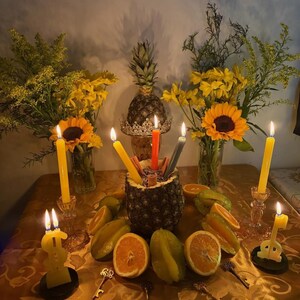 Pu&ograve; includere: Un centrotavola con un ananas svuotato riempito con tre candele affusolate accese nei colori rosso, giallo e grigio. Attorno all'ananas ci sono fette d'arancia, carambole e quattro piccole chiavi di metallo. Due vasi di fiori gialli e girasoli fiancheggiano la composizione, con ulteriori candele gialle accese e candele a forma di simbolo del dollaro.