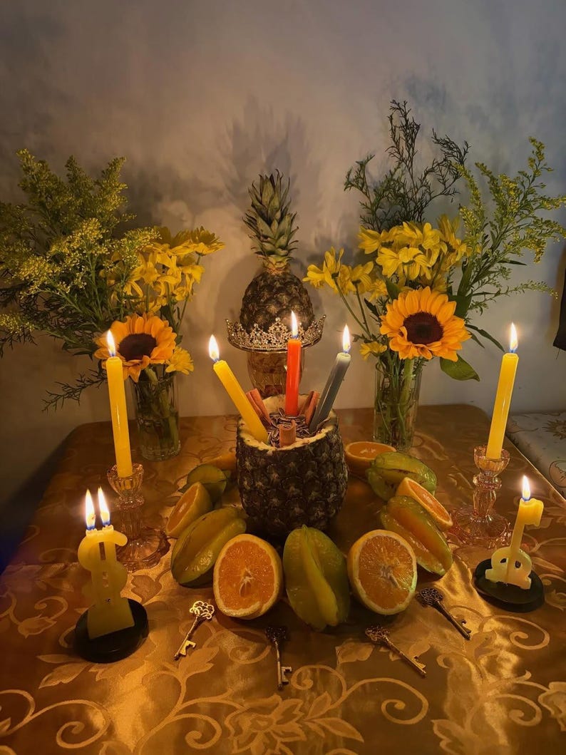 Pu&ograve; includere: Un centrotavola con un ananas svuotato riempito con tre candele affusolate accese nei colori rosso, giallo e grigio. Attorno all'ananas ci sono fette d'arancia, carambole e quattro piccole chiavi di metallo. Due vasi di fiori gialli e girasoli fiancheggiano la composizione, con ulteriori candele gialle accese e candele a forma di simbolo del dollaro.