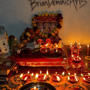 Pu&ograve; includere: Un altare ind&ugrave; con un'immagine centrale di una divinit&agrave; incorniciata, decorata con ghirlande di calendule gialle e rosse. Diverse lampade a olio in terracotta accese, chiamate diya, sono disposte su un panno rosso e sul pavimento. Il testo BrianDemonicArts &egrave; visibile.