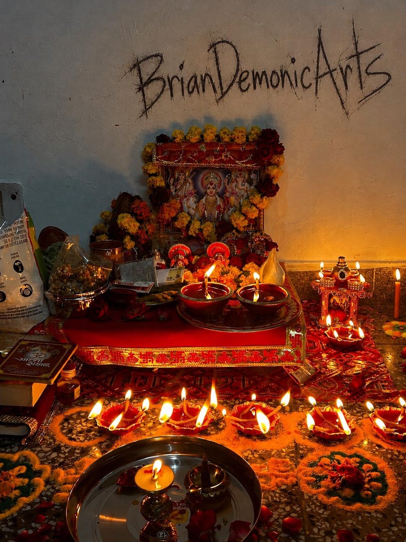 Pu&ograve; includere: Un altare ind&ugrave; con un'immagine centrale di una divinit&agrave; incorniciata, decorata con ghirlande di calendule gialle e rosse. Diverse lampade a olio in terracotta accese, chiamate diya, sono disposte su un panno rosso e sul pavimento. Il testo BrianDemonicArts &egrave; visibile.