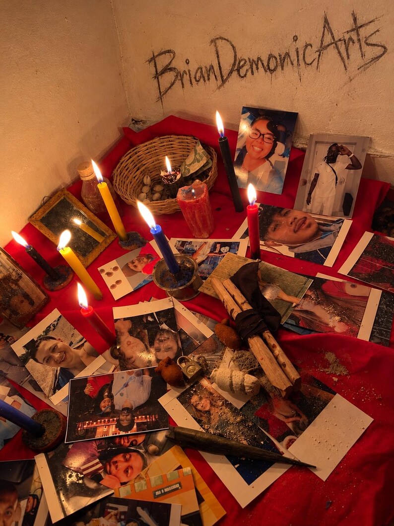 May include: A red cloth surface covered with various photographs of people, multiple lit candles in yellow, blue, red, and black, a woven basket containing small items, a small mirror, and a wooden bundle. The text BrianDemonicArts is written on the wall.