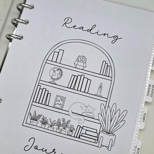 Könnte beinhalten: Eine weiße Reading Journal-Seite mit einer schwarzen Strichzeichnung eines Bücherregals, eines Globus, Pflanzen und einer schlafenden Katze. Das Wort "Reading" steht über dem Bücherregal und "Journal" darunter. Das Journal befindet sich in einem weißen Ordner.