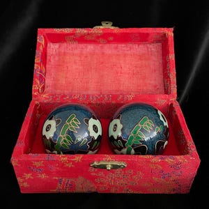 Può includere: Due sfere cinesi per la salute blu con disegni floreali bianchi e accenti verdi, all'interno di una scatola rossa foderata in tessuto. La scatola ha una chiusura color oro ed è posta su uno sfondo nero.