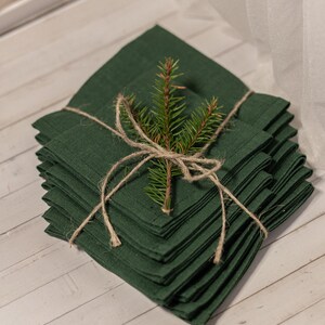 Puede incluir: Una pila de servilletas de lino verde oscuro atadas con hilo y una ramita de hoja perenne. Las servilletas están cuidadosamente dobladas y dispuestas sobre una superficie de madera clara, creando una presentación rústica y festiva.
