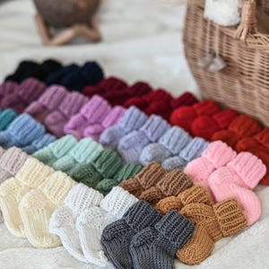 Peut inclure: Une collection de chaussons de bébé tricotés colorés, dans des tons variés, dont blanc, gris, rose et bleu. Les chaussons sont disposés en rangées sur une surface blanche et douce, avec un panier en osier et des éléments décoratifs en arrière-plan.