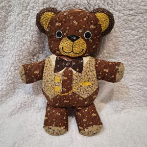 Puede incluir: Un oso de peluche hecho a mano con un estampado floral marrón. El oso tiene un chaleco amarillo y marrón con una pajarita y un diseño de reloj de bolsillo. Los ojos del oso son círculos negros, y la nariz y la boca son amarillas.