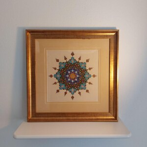 May include: A framed artwork featuring a detailed, symmetrical design in shades of blue, teal, orange, and brown. The artwork is set against a cream-colored background and enclosed in a gold-colored wooden frame. The artwork is displayed on a white shelf against a light blue wall.