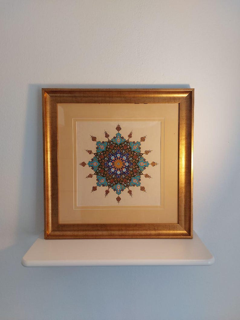 May include: A framed artwork featuring a detailed, symmetrical design in shades of blue, teal, orange, and brown. The artwork is set against a cream-colored background and enclosed in a gold-colored wooden frame. The artwork is displayed on a white shelf against a light blue wall.