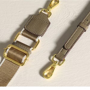 May include: Close-up of a tan and white striped bag strap with gold-colored metal hardware. The strap has a leather accent and a gold-colored clasp. The strap is designed to be attached to a purse or handbag.