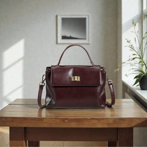 Peut inclure: Un sac à main en cuir bordeaux avec une poignée supérieure et une bandoulière amovible. Le sac a une forme rectangulaire avec une fermeture à rabat et un fermoir doré. Le sac est posé sur une table en bois.