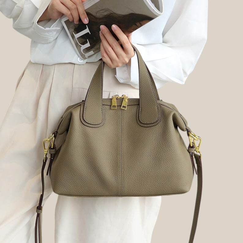 May include: A taupe-colored handbag with a textured finish. The bag features two top handles and a detachable shoulder strap. Gold-toned zippers and hardware add a touch of elegance to the design. The bag is held by a person wearing a white shirt.