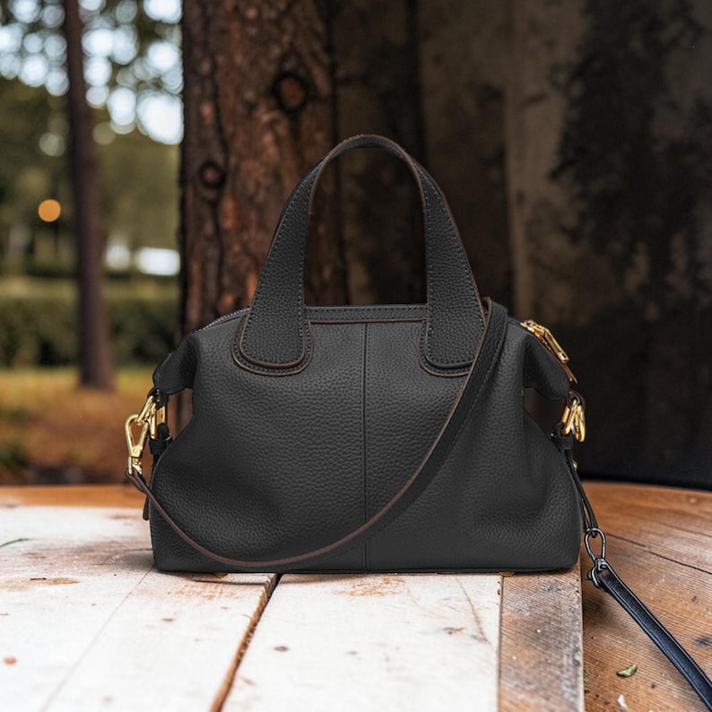 May include: Black leather handbag with a top handle and a detachable shoulder strap. The bag has gold-colored hardware and a textured surface. The bag is sitting on a wooden surface.