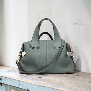 May include: A sage green leather handbag with a top handle and a detachable shoulder strap. The bag features gold-colored hardware and a structured design. The bag is sitting on a wooden surface.