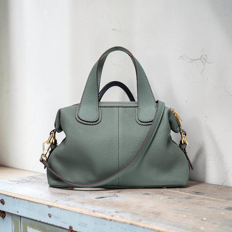 May include: A sage green leather handbag with a top handle and a detachable shoulder strap. The bag features gold-colored hardware and a structured design. The bag is sitting on a wooden surface.
