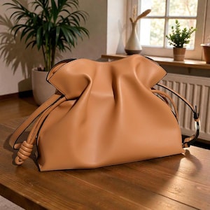 Peut inclure: Un sac à main en cuir beige avec une fermeture à cordon et des détails noués. Le sac a un design souple et froncé et de longues sangles. Il est posé sur une surface en bois, avec un arrière-plan flou avec des plantes et une fenêtre.
