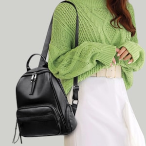 Puede incluir: Una mochila de cuero negro con la parte superior redondeada, bolsillo frontal y cremalleras plateadas. La mochila tiene correas ajustables y un asa superior. La bolsa se muestra sobre un fondo claro.