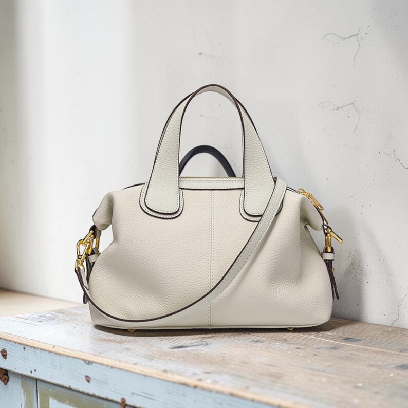 May include: Cream-colored leather handbag with a structured shape. Features a top handle and a detachable shoulder strap. Gold-toned hardware accents the bag, which is approximately 12 inches wide.
