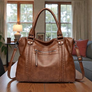 Peut inclure: Un sac fourre-tout en cuir marron avec une poignée supérieure et une bandoulière amovible. Le sac comporte une poche avant zippée, des accents latéraux tressés et une quincaillerie argentée. Le sac est sur une table en bois.