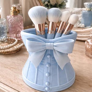 May include: A light blue corset-shaped makeup brush holder with a large bow and pearl-like buttons. The holder contains four makeup brushes with light blue bristles and rose gold metal handles. The holder is on a wooden surface.