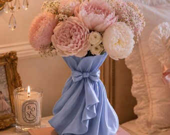 May include: A light blue vase with a sculpted fabric-like texture and a small bow detail at the neck, filled with a bouquet of pink and white peonies and baby's breath. The vase rests on two stacked books titled Dior Catwalk and Chanel.
