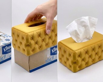 May include: A rectangular mustard yellow tissue box cover featuring a tufted button design. The cover is shown being placed over a standard cardboard Kleenex tissue box, resulting in a finished, upholstered-look tissue dispenser.