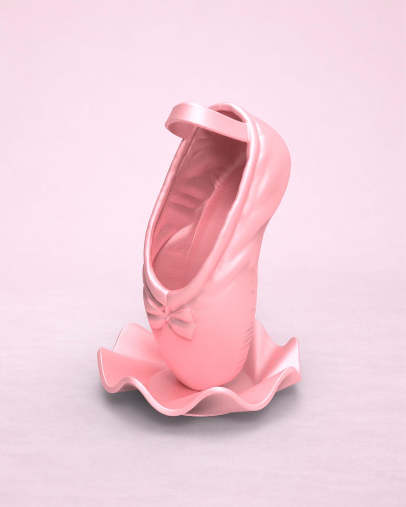May include: A light pink ballet slipper sculpture standing upright on a matching wavy, circular base. The shoe features a small decorative bow at the toe and a single strap across the top opening, all rendered in a smooth, solid matte finish.