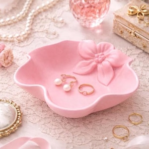 May include: A pink ceramic jewelry dish with a flower design and two rings. The dish holds a pair of pearl earrings and a gold ring with a pink stone. Other jewelry and accessories are in the background.
