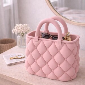 May include: A pink handbag with a quilted design, featuring two rounded handles. The bag contains sunglasses and keys. It sits on a light-colored wooden surface, with a mirror and flowers in the background.