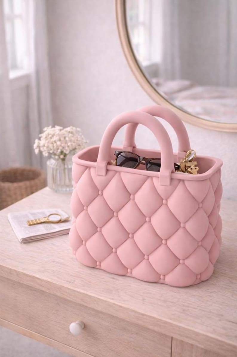 May include: A pink handbag with a quilted design, featuring two rounded handles. The bag contains sunglasses and keys. It sits on a light-colored wooden surface, with a mirror and flowers in the background.