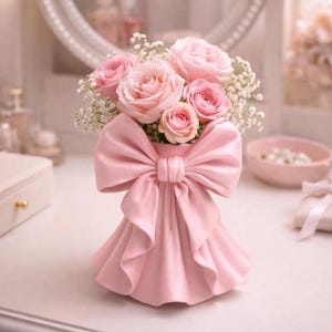 Könnte beinhalten: Eine rosa dekorative Vase in Form eines Kleides mit einer großen Schleife, die einen Strauß rosa Rosen und weiße Füllblumen hält. Die Vase steht auf einer weißen Oberfläche, mit einem Spiegel und anderen dekorativen Gegenständen im Hintergrund.