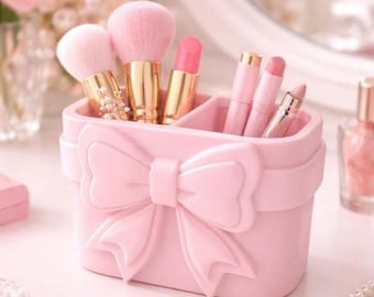 May include: A pink makeup organizer with a bow design, holding pink makeup brushes and lipsticks. The organizer is on a white surface, with a mirror and other beauty products in the background.