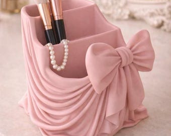 May include: A pink makeup brush holder with a draped fabric design and a large bow. The holder contains two makeup brushes and a pearl necklace. The holder is made of a solid material and is sitting on a light-colored surface.