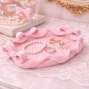 May include: A light pink decorative tray with bow accents, holding a pearl bracelet, rings, and a snowflake-shaped pendant. Perfume bottles and a gold-framed mirror are in the background, creating a feminine aesthetic.