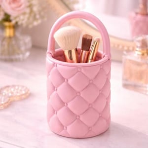 May include: A pink, quilted makeup brush holder with a handle, containing several makeup brushes. The holder is cylindrical and features a diamond pattern. The brushes have gold-coloured handles and various brush heads. The background includes a mirror and perfume bottles.