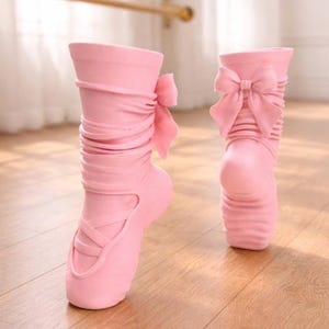 May include: Two pink ballet slipper-shaped planters. Each planter has a bow on the side and a crisscross design on the front. The planters are made of a smooth material and sit on a wooden floor.